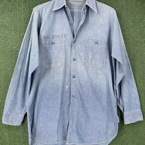 May include: Light blue denim shirt with long sleeves, two chest pockets, and button closures. The text "NELSON S.O." is embroidered on the left chest pocket. The shirt is hanging on a wooden hanger.