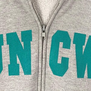 May include: A close-up of a grey zip-up hoodie featuring the teal letters "UNCW". The hoodie has a full zip and a soft, fleecy interior. The letters are in a bold, blocky font.