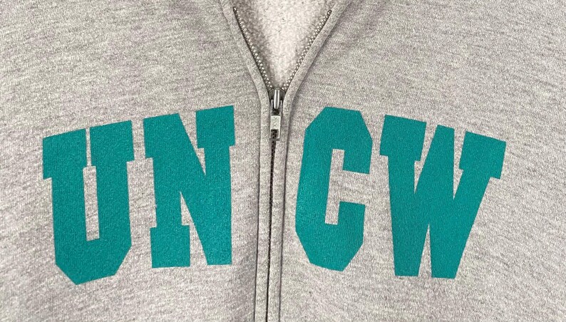 May include: A close-up of a grey zip-up hoodie featuring the teal letters "UNCW". The hoodie has a full zip and a soft, fleecy interior. The letters are in a bold, blocky font.