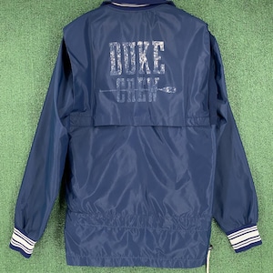 Puede incluir: Chaqueta azul marino con puños a rayas blancas y azul marino. La parte trasera de la chaqueta muestra el texto "DUKE CREW" en plata desgastado, con un gráfico de remo. La chaqueta tiene un cordón blanco y un cuello azul marino.