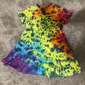 May include: A tie-dye dress in a rainbow of colors, including red, orange, yellow, green, blue, and purple. The dress has short sleeves and a tiered skirt. The fabric has a swirling, mottled pattern.