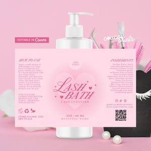 Puede incluir: Una botella rosa y blanca de limpiador de pestañas Lash Bath con un diseño de corazón. La etiqueta incluye instrucciones e ingredientes. Un recipiente negro con cepillos de pestañas rosas y perlas en el fondo.