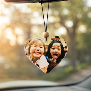 May include: A heart-shaped car air freshener or hanging ornament featuring a color photograph of two children smiling and laughing. The ornament is suspended by a thin black cord with a small, round, light-colored wooden bead at the top.