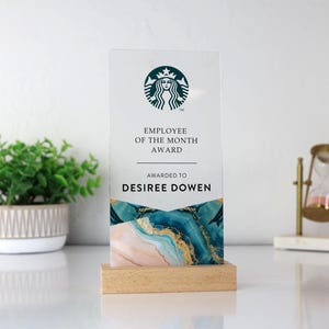 May include: A clear acrylic Employee of the Month award with the Starbucks logo at the top. The award features the text "Employee of the Month Award" and "Awarded to Desiree Dowen". The base is made of light-colored wood, and the bottom of the award has a blue, gold, and pink design.