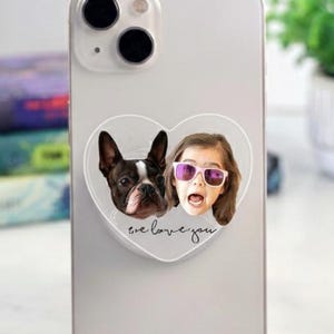 May include: A silver smartphone with a heart-shaped phone grip featuring a Boston Terrier and a girl wearing sunglasses. The grip has the text "we love you". A clear, oval-shaped phone grip with a woman's face is also visible.