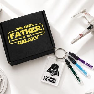 May include: A black gift box with the text "THE BEST FATHER IN THE GALAXY" in yellow. A keychain with a Darth Vader graphic and the text "I AM THEIR FATHER" is attached to three personalized keychains with names. A Star Wars themed gift.