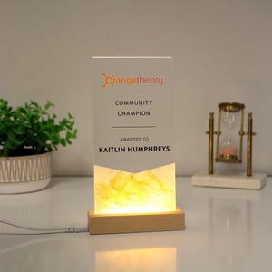 May include: A rectangular acrylic award with the Orangetheory logo and the text "COMMUNITY CHAMPION AWARDED TO KAITLIN HUMPHREYS." The award is illuminated from the base with a warm yellow light, and sits on a wooden base.