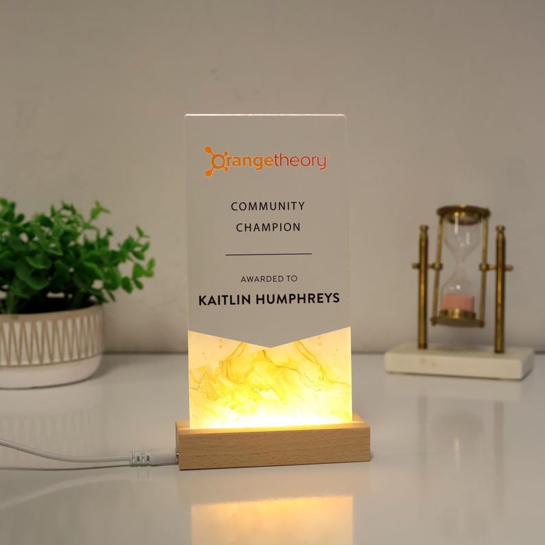 May include: A rectangular acrylic award with the Orangetheory logo and the text "COMMUNITY CHAMPION AWARDED TO KAITLIN HUMPHREYS." The award is illuminated from the base with a warm yellow light, and sits on a wooden base.