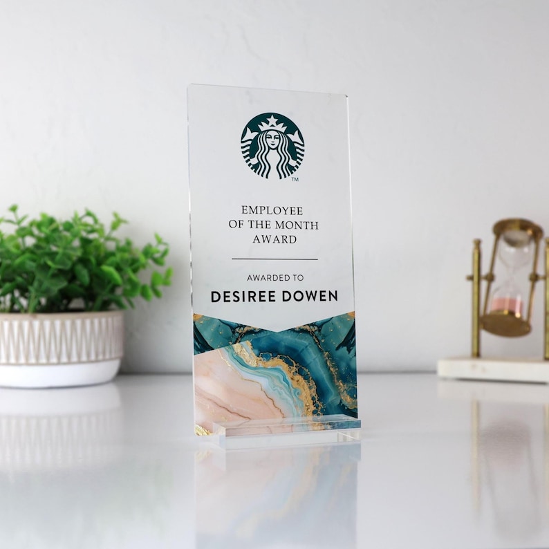 May include: A clear acrylic Employee of the Month award featuring the Starbucks logo and the text "Employee of the Month Award" and "Awarded to Desiree Dowen." The bottom of the award has a design with blue, gold, and pink marble-like patterns.