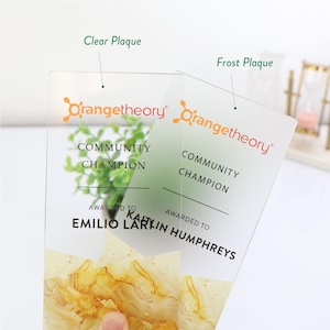May include: Two clear and frosted acrylic plaques with the Orangetheory logo and the words "Community Champion." The plaques are personalized with names and feature a gold and yellow marbled design at the bottom.
