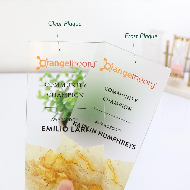 May include: Two clear and frosted acrylic plaques with the Orangetheory logo and the words "Community Champion." The plaques are personalized with names and feature a gold and yellow marbled design at the bottom.