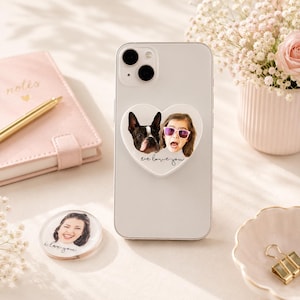 May include: A white smartphone with a heart-shaped phone grip featuring a photo of a dog and a girl with the text "we love you." A clear phone grip with a photo of a woman and the text "i love you" is also visible.