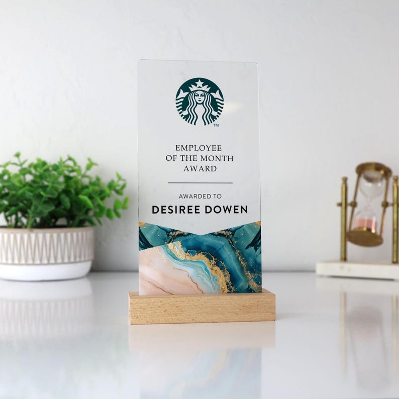 May include: A clear acrylic Employee of the Month award with the Starbucks logo at the top. The award features the text "Employee of the Month Award" and "Awarded to Desiree Dowen". The base is made of light-colored wood, and the bottom of the award has a blue, gold, and pink design.