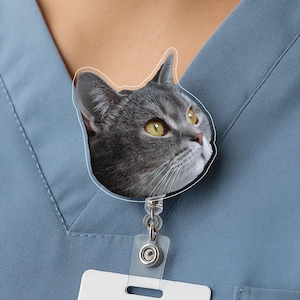 May include: A gray cat face badge reel with yellow eyes. The cat's head is outlined in white and attached to a clear plastic reel with a white rectangular tag. The badge reel is clipped to a blue top.