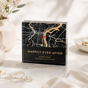 May include: A black and gold map design on a clear acrylic block. The map features a red heart and the words "HAPPILY EVER AFTER" with a couple's names and coordinates. A decorative wedding or anniversary gift.
