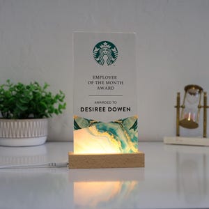 May include: An illuminated acrylic Employee of the Month award. The award features the Starbucks logo, the text "Employee of the Month Award," and the name "Desiree Dowen." The base is wood, and the design includes teal and gold accents.