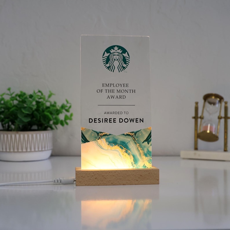 May include: An illuminated acrylic Employee of the Month award. The award features the Starbucks logo, the text "Employee of the Month Award," and the name "Desiree Dowen." The base is wood, and the design includes teal and gold accents.
