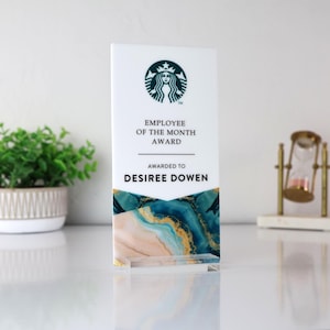 May include: A white acrylic award with the Starbucks logo and the text "EMPLOYEE OF THE MONTH AWARD AWARDED TO DESIREE DOWEN." The bottom of the award features a blue, gold, and peach marbled design. The award is displayed on a clear acrylic stand.