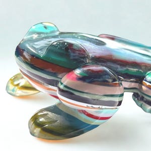 Peut inclure: Une sculpture de grenouille en verre multicolore. La grenouille est faite de couches de verre translucide dans les tons de bleu, vert, rouge et marron. La grenouille a un corps lisse et arrondi et quatre petites pattes.