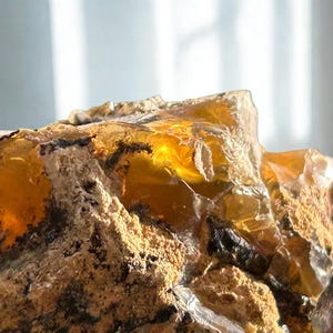 Op de afbeelding: Close-up van een ruwe, amberkleurige edelsteen met een kristallijne structuur. De steen heeft een mix van goudgele en bruine tinten, met gebieden van doorschijnendheid. Het oppervlak is getextureerd en ongelijkmatig, met zichtbare insluitsels en onvolkomenheden.