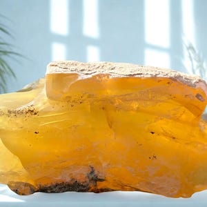 Puede incluir: Una gran gema de color ámbar con una superficie rugosa y texturizada. La piedra tiene una cualidad translúcida, permitiendo el paso de la luz, y se encuentra sobre un fondo suave y neutro. Los tonos cálidos y la forma única de la gema la convierten en un objeto decorativo llamativo.
