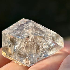 Op de afbeelding: Een helder kwartskristal met een facetvormige, geometrische vorm. Het kristal heeft een enigszins ruw, getextureerd oppervlak en een natuurlijke, aardse basis. Het kristal wordt in een hand gehouden, met een donkere achtergrond.