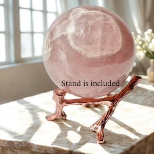 Peut inclure: Une grande sphère en quartz rose poli avec des inclusions naturelles et une teinte rose pâle. La sphère repose sur un support métallique de couleur cuivre en forme de branche d'arbre. Le texte "Stand is included" est inscrit sur la sphère.