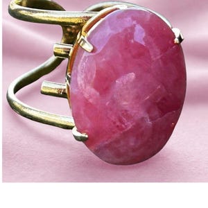 May include: A gold ring with a large, oval pink gemstone. The gemstone is set in a gold bezel, and the ring has an open design. The background is a soft pink fabric, creating a contrast with the ring.