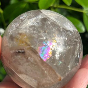 May include: A clear, spherical quartz crystal ball featuring internal fractures and inclusions. The stone displays a vibrant, multi-colored rainbow refraction effect within its polished surface, highlighting natural mineral patterns and light play.
