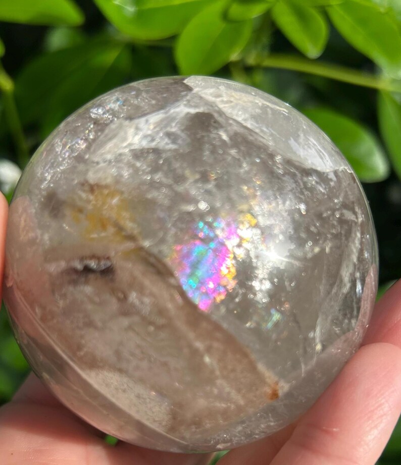 May include: A clear, spherical quartz crystal ball featuring internal fractures and inclusions. The stone displays a vibrant, multi-colored rainbow refraction effect within its polished surface, highlighting natural mineral patterns and light play.