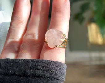 Puede incluir: Un anillo de oro con una gran piedra de cuarzo rosa de color rosa claro. El anillo se lleva en un dedo.