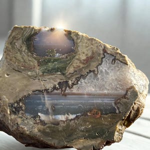 May include: A natural stone specimen with a variety of colours. The stone features areas of brown, green, and white, with a translucent, crystalline section. The overall shape is irregular, showcasing the raw, unpolished nature of the material.