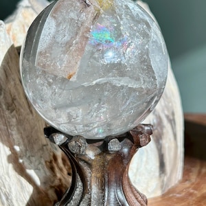 May include: A clear quartz crystal sphere featuring internal fractures, cloudy inclusions, and a small rainbow refraction. The sphere is supported by a dark brown, carved wooden stand with a textured, organic shape and a flared base.