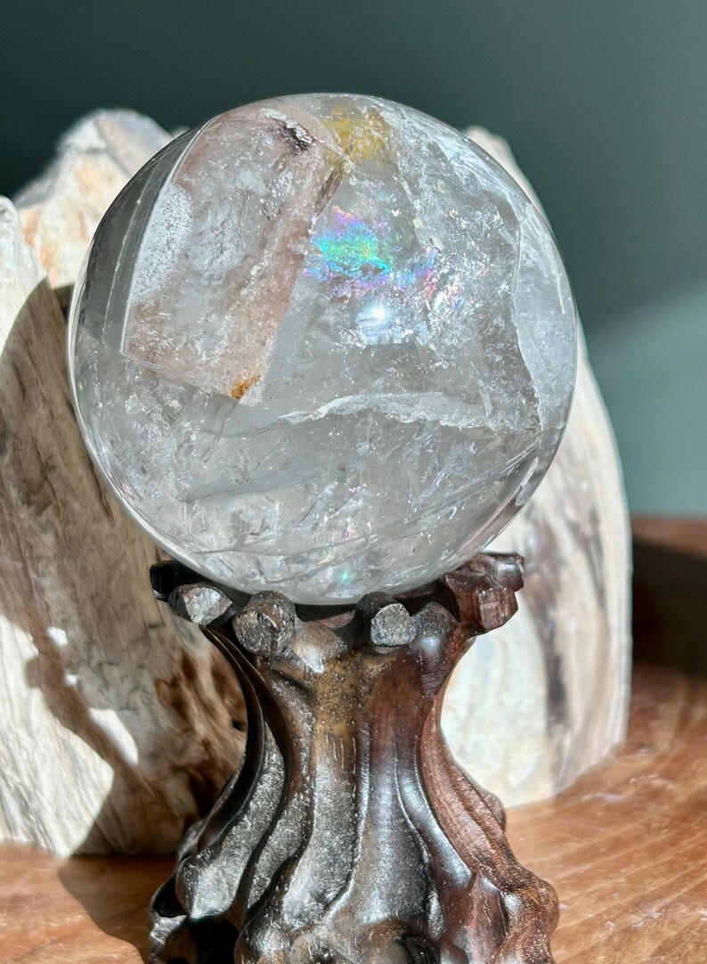 May include: A clear quartz crystal sphere featuring internal fractures, cloudy inclusions, and a small rainbow refraction. The sphere is supported by a dark brown, carved wooden stand with a textured, organic shape and a flared base.