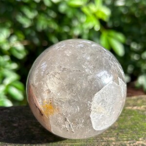 May include: A clear, spherical quartz crystal ball featuring internal fractures, natural inclusions, and a small patch of yellow mineral deposits. The surface is smooth and polished, reflecting light across its rounded, transparent structure.