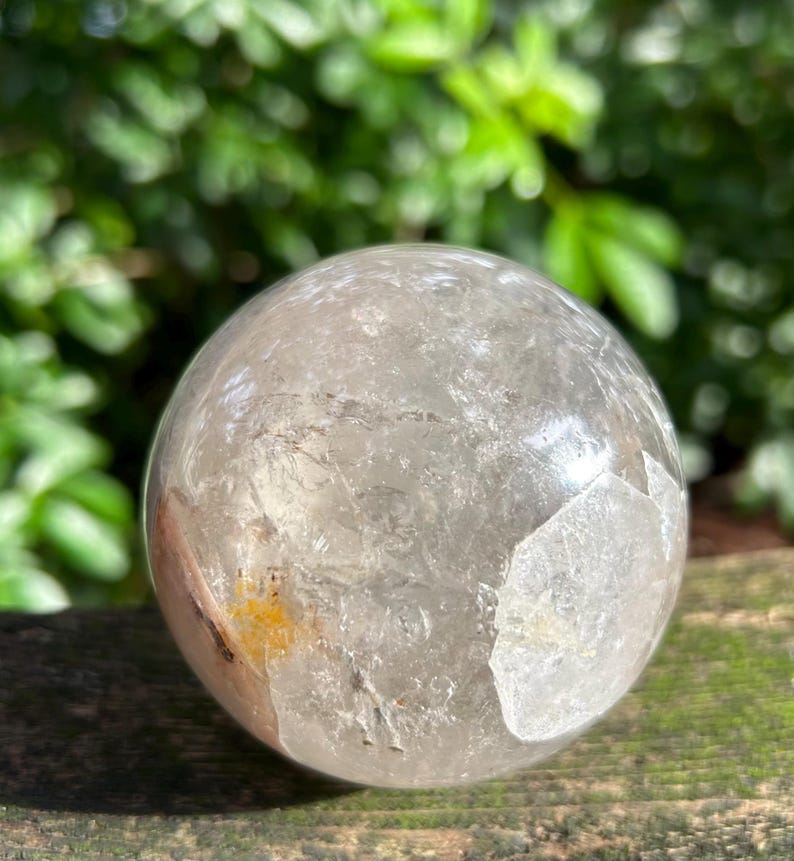May include: A clear, spherical quartz crystal ball featuring internal fractures, natural inclusions, and a small patch of yellow mineral deposits. The surface is smooth and polished, reflecting light across its rounded, transparent structure.