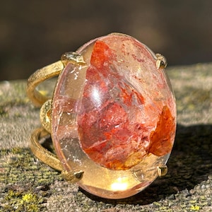 May include: A gold ring with a large, oval gemstone. The gemstone has red and orange inclusions. The ring's band has a textured design. The ring is displayed on a textured surface. The ring's design is eye-catching.