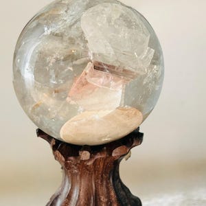 May include: A clear, spherical quartz crystal ball featuring internal mineral inclusions and a distinct, opaque tan-colored patch. The sphere is supported by a dark brown, carved wooden stand with a fluted, pedestal-style base design.