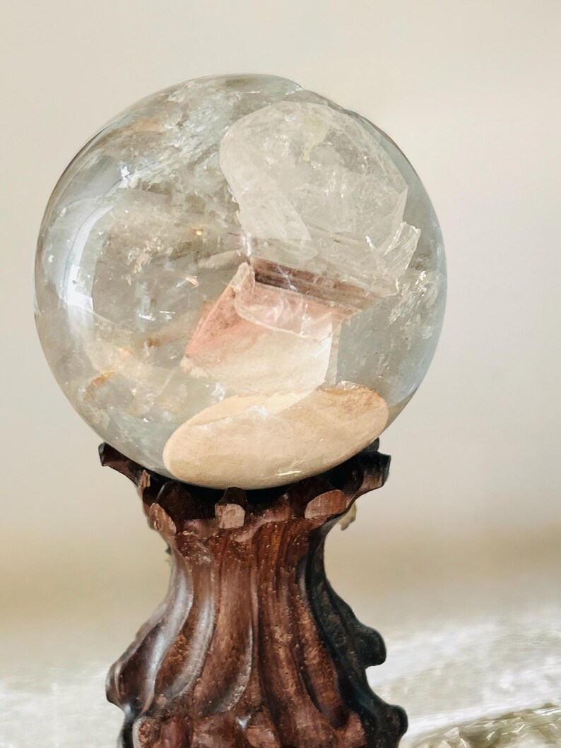 May include: A clear, spherical quartz crystal ball featuring internal mineral inclusions and a distinct, opaque tan-colored patch. The sphere is supported by a dark brown, carved wooden stand with a fluted, pedestal-style base design.