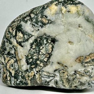 Könnte beinhalten: Ein Naturstein mit einer Mischung aus Farben, darunter Weiß, Schwarz und Beige. Der Stein hat eine glatte, abgerundete Form und ein einzigartiges Muster aus Adern und Texturen. Das Gesamtbild ist erdig und organisch.