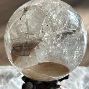 May include: A clear, spherical quartz crystal ball featuring internal fractures, natural inclusions, and a distinct tan-colored sediment layer at the base. The polished surface reflects light, highlighting the complex, multi-faceted internal structure.