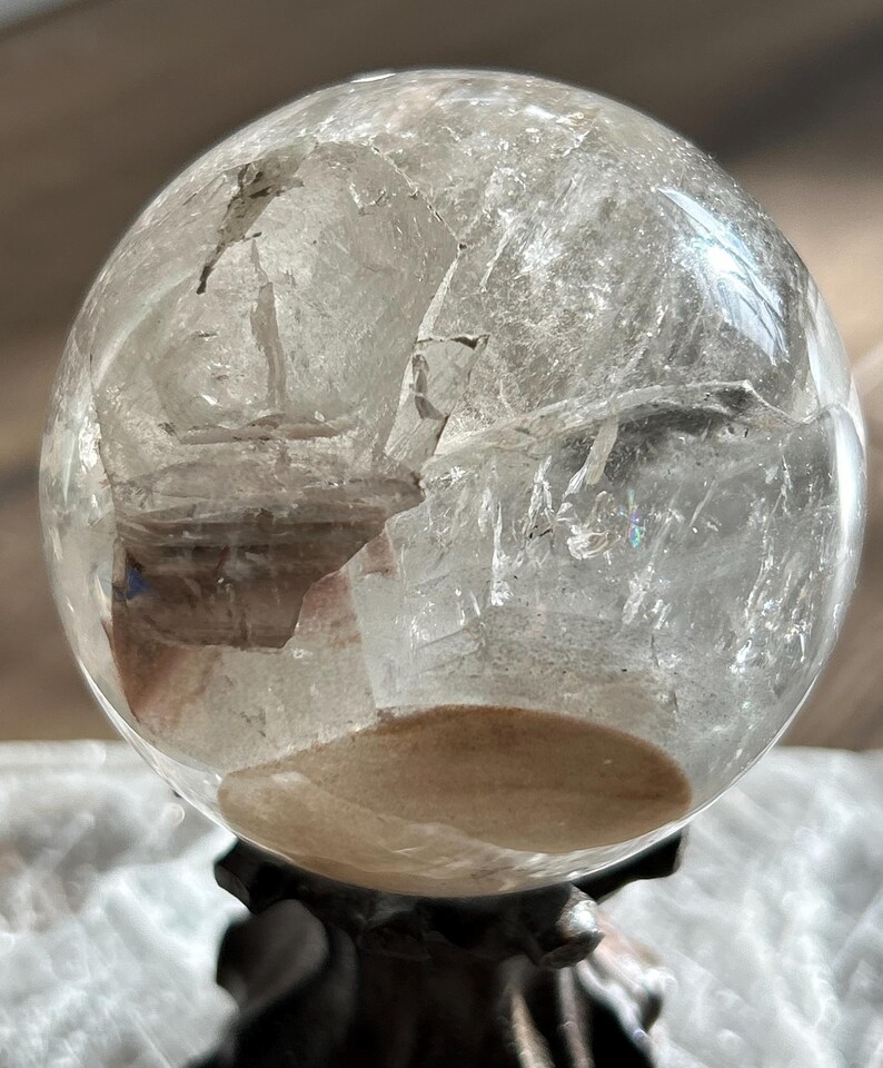 May include: A clear, spherical quartz crystal ball featuring internal fractures, natural inclusions, and a distinct tan-colored sediment layer at the base. The polished surface reflects light, highlighting the complex, multi-faceted internal structure.
