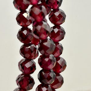 May include: Close-up of strands of faceted, deep red garnet beads. The beads are strung together, showcasing their polished, reflective surfaces. The gemstones are a rich, dark red colour, with visible facets catching the light.