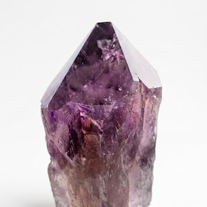 以下が含まれることがあります： 紫色の大きなアメジストの結晶。結晶は、より明るい色合いと自然な質感を持つ深い紫色をしています。上部は鋭い幾何学的なポイントで、基部は粗く未研磨です。