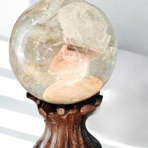 May include: A clear, spherical quartz crystal ball featuring internal mineral inclusions and a smooth, polished surface. The sphere rests securely on a dark brown, carved wooden stand with a fluted, pedestal-style base and scalloped edges.