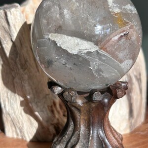 May include: A spherical clear quartz crystal ball featuring internal white mineral inclusions and a subtle golden-brown tint. The polished stone rests securely on a dark brown, carved wooden stand with a textured, organic, multi-pronged base.