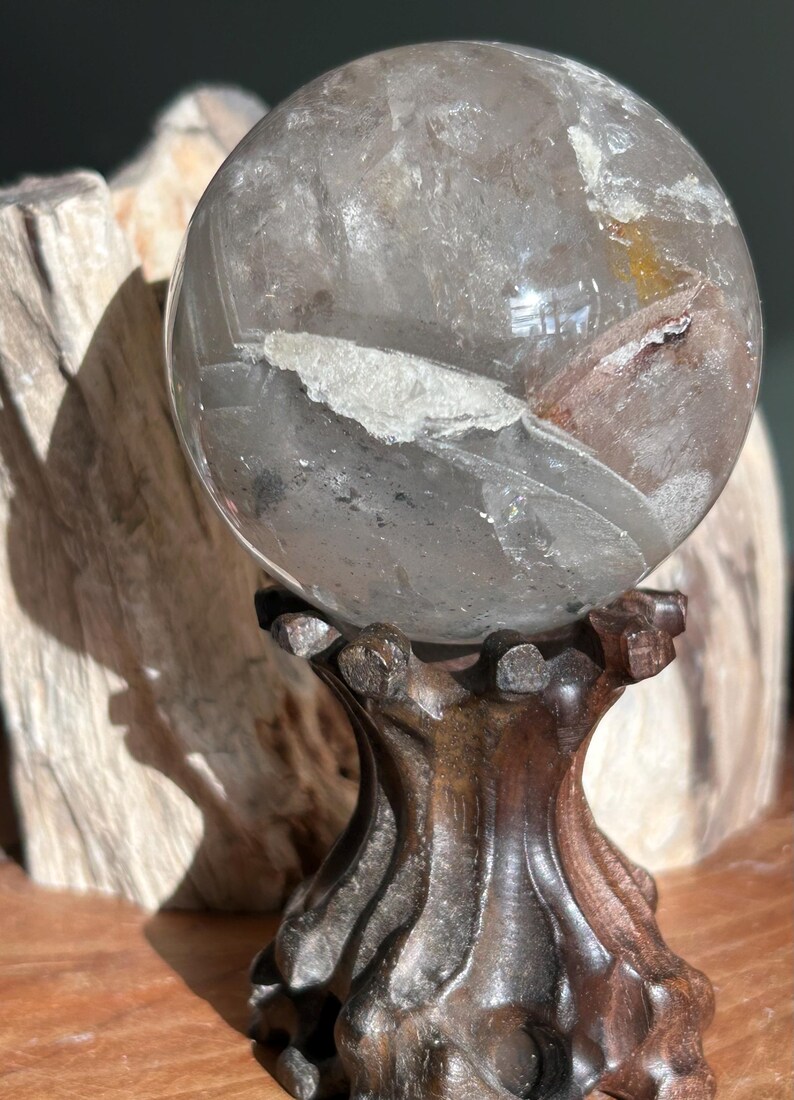 May include: A spherical clear quartz crystal ball featuring internal white mineral inclusions and a subtle golden-brown tint. The polished stone rests securely on a dark brown, carved wooden stand with a textured, organic, multi-pronged base.