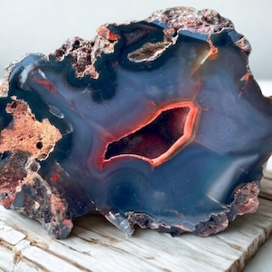Puede incluir: Una rodaja de geoda de ágata pulida que presenta un centro azul profundo y gris translúcido con una vibrante cavidad cristalina de color naranja rojizo. El borde exterior de la piedra está compuesto por una corteza mineral rugosa, texturizada, de color marrón oscuro y marrón rojizo.