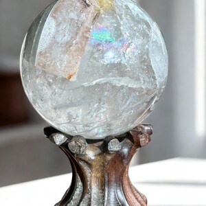 May include: A clear, spherical quartz crystal ball featuring internal fractures and a visible rainbow refraction, resting securely on a dark, carved wooden stand with a textured, organic shape and a polished finish.