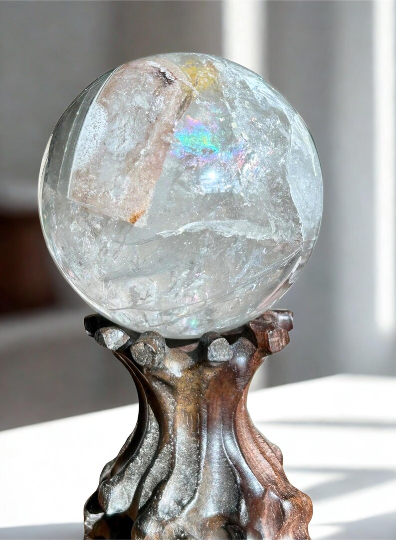 May include: A clear, spherical quartz crystal ball featuring internal fractures and a visible rainbow refraction, resting securely on a dark, carved wooden stand with a textured, organic shape and a polished finish.